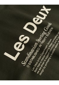 Tmavozelená tkanina s bielym textom, prezentujúca značku "Les Deux", s ďalšími detailmi o založení a dizajnovej filozofii.