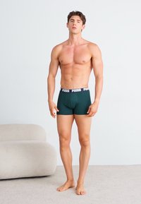 Boxer briefs verde-escuro com uma faixa na cintura com o logótipo da Puma em preto e branco, modelo a pé descalço, fundo apresenta um sofá de cor clara.