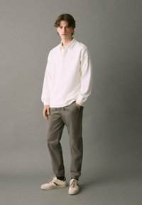 Ung man står med händerna i fickorna och har på sig en vit långärmad polotröja, grå joggbyxor och vita sneakers med gröna ränder.