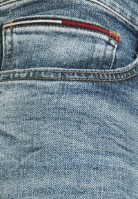 Tissu en denim bleu clair montrant un gros plan d'une poche arrière avec des bandes décoratives rouges, blanches et bleues, et des coutures contrastantes jaunes.