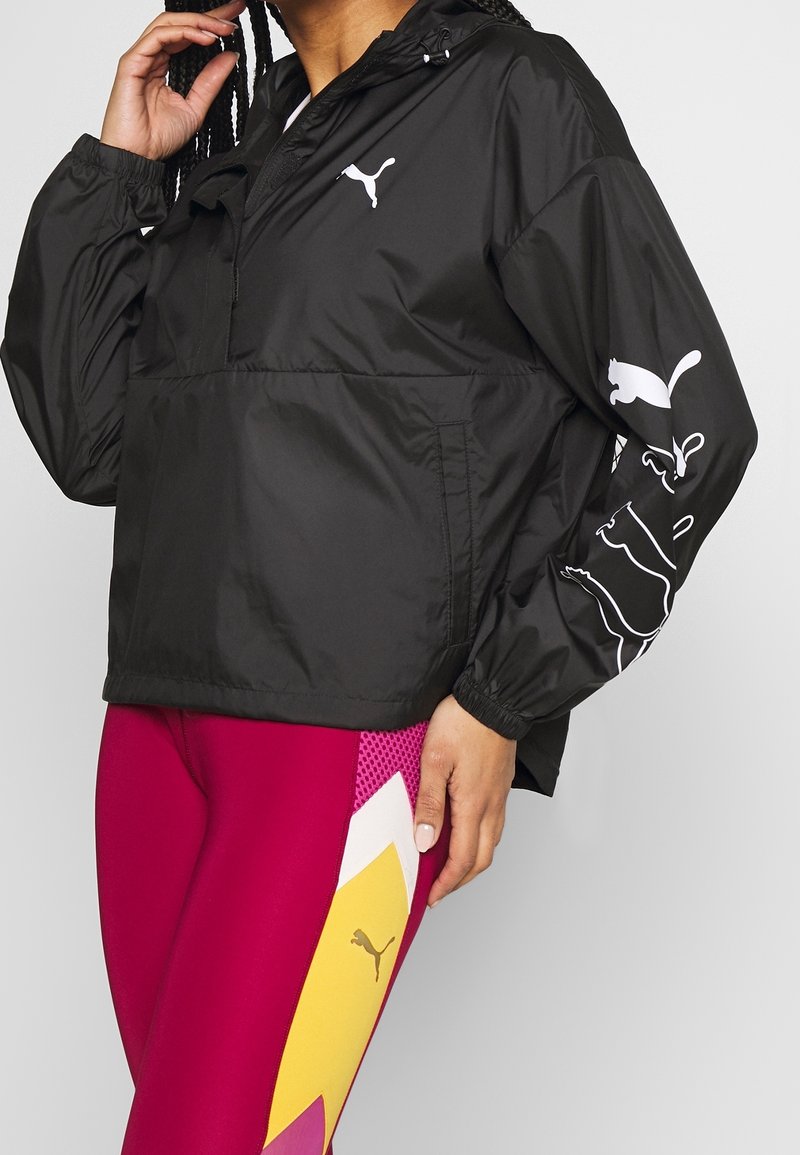 Personne portant un coupe-vent noir Puma avec un logo blanc, associé à des leggings magenta avec des motifs géométriques jaunes et blancs et le logo Puma.