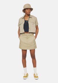 Camicia cropped beige con bottoni e gonna abbinata, abbinata a una canotta scura e sneakers gialle. Presenta accenti di logo sottili e tasche.