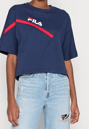 Persoon draagt een marineblauw FILA crop top met een rode diagonale streep en lichtblauwe gescheurde jeans tegen een effen achtergrond.