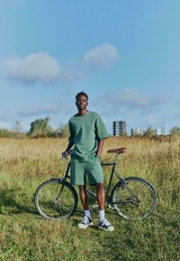Tenue composée d'un short vert et d'un t-shirt surdimensionné, associée à un vélo noir avec une selle en cuir marron, installée dans un champ herbeux sous un ciel bleu.