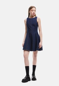 Donkerblauwe denim A-lijn jurk met mouwloos ontwerp, zichtbare naden en een uitlopende rok. Gecombineerd met zwarte plateauschoenen en sokken.