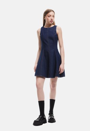 Donkerblauwe denim A-lijn jurk met mouwloos ontwerp, zichtbare naden en een uitlopende rok. Gecombineerd met zwarte plateauschoenen en sokken.