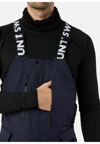 Marineblauwe bretels met verstelbare banden, ritszakken en een strakke pasvorm. Draagt over een zwart geribbeld longsleeve top.