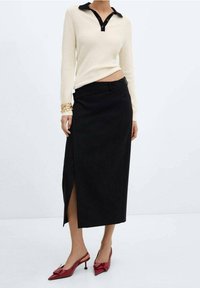 Jupe noire longue avec une fente sur le côté, associée à un pull côtelé crème avec un col noir et des détails de boutons, portée avec des escarpins rouges �à bout pointu.