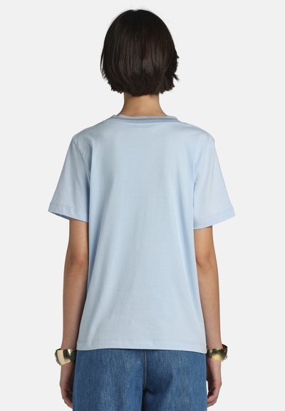 Person med kort mørkt hår iført en lyseblå kortærmet skjorte og blå jeans, vist bagfra på en hvid baggrund.