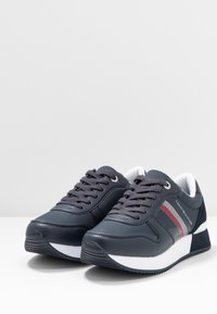 Granatowe syntetyczne sneakersy o gładkiej teksturze, z metalowymi oczkami, białymi akcentami i czerwonym paskiem na boku.