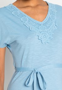 Světle modré šaty s výstřihem do V a jemnými krajkovými detaily, krátkými rukávy a textilním opaskem uvázaným v pase. Měkká textura.