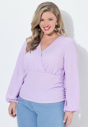 Glimlachende vrouw met lang blond haar die grote oorringen draagt, een gestructureerde lavendelkleurige V-hals blouse en lichtblauwe spijkerbroek tegen een witte achtergrond.