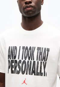 Κοντινό πλάνο ενός ατόμου που φοράει λευκό πουκάμισο με μεγάλα μαύρα γράμματα: «ΚΑΙ ΤΟ ΠΗΡΑ ΠΡΟΣΩΠΙΚΑ» και ένα μικρό κόκκινο λογότυπο Jumpman από κάτω.