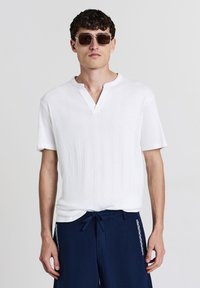 Uomo che indossa occhiali da sole scuri, una camicia bianca a maniche corte con texture e colletto a V, e pantaloni blu navy con cordoncino e motivi laterali.