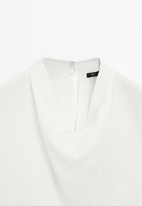Blouse blanche à encolure drapée avec petite fermeture à bouton au dos du col, montrant à l’intérieur une étiquette noire de la marque.