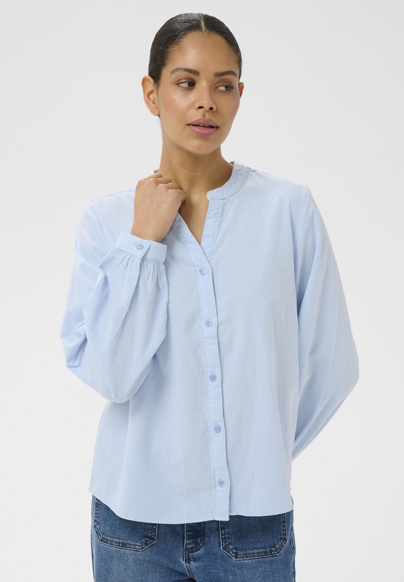 Femme aux cheveux foncés portant un chemisier bleu clair à manches longues boutonné, debout devant un fond clair uni, regardant sur le côté.