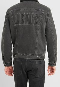 Henkilö, joka on pukeutunut mustaan Levi's-denim-takkiin, jossa on iso kohokuvioitu logo takana, ja mustiin Wrangler-farkkuihin, näkyy takaa.