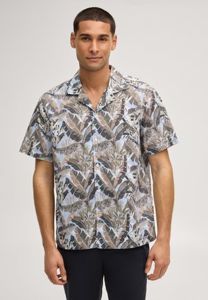 Mann mit kurzen Haaren, der ein kurzärmeliges Hemd mit braunem und grünem Tropenblattmuster auf hellblauem Hintergrund trägt.