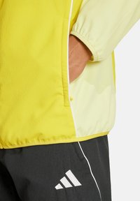 Gelbe, leichte Jacke mit strukturierter Oberfläche, ausgestattet mit einer Seitentasche und weißen Akzenten. Getragen über dunklen Hosen mit dem Adidas-Logo.