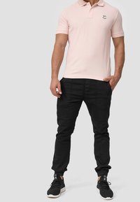 Mężczyzna w jasnoróżowej koszulce polo, czarnych spodniach dresowych i czarnych sneakersach Adidas stoi z jedną ręką w kieszeni.