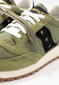 Oliivinvihreät urheilukengät, joissa on yhdistettyä tekstiilisuedea ja nailonia, valkoiset nauhat, mustat yksityiskohdat ja kevyt kumipohja, jossa on uurretut kuviot.