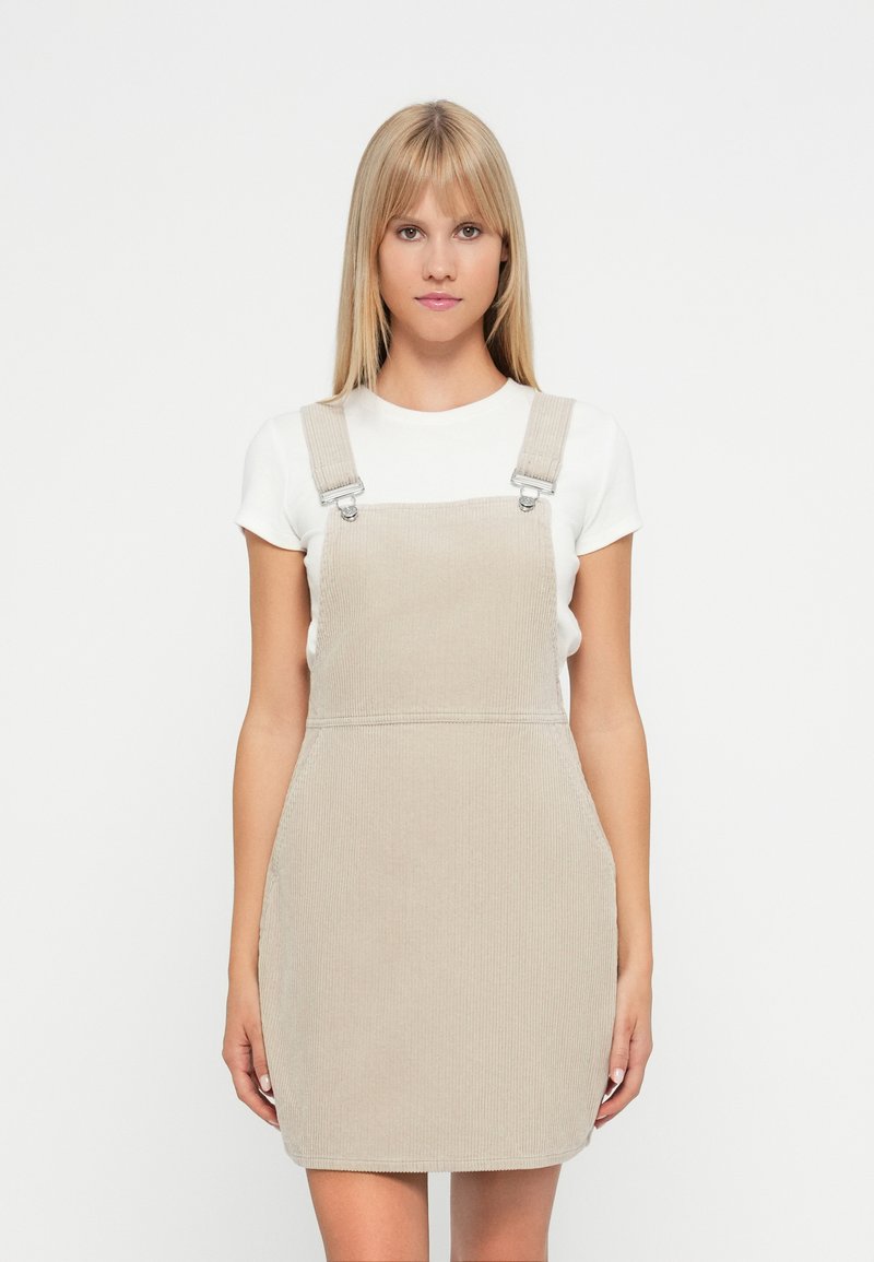 Robe salopette en corduroy beige avec bretelles réglables et poches latérales, présentant une silhouette ajustée et une texture côtelée.