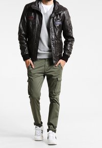 Schwarze Leder-Bomberjacke mit Patches, grauer Pullover, olivgrüne Cargo-Hose und weiße Sportschuhe. Glatte Textur mit gerippten Bündchen.