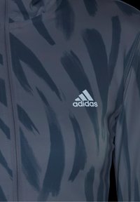 Veste de sport bleu foncé avec un motif de coups de pinceau noir et un logo Adidas blanc sur la poitrine, avec une fermeture éclaire à l'avant.