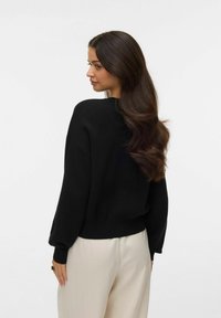 Suéter negro de punto con un ajuste relajado, hombros caídos y puños acanalados, combinado con pantalones de color claro. Textura suave y diseño sencillo.