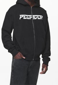 Sudadera con cremallera negra de algodón, con un gran logo blanco de "PEGADOR" en el pecho, bolsillos frontales y puños y dobladillo acanalados.
