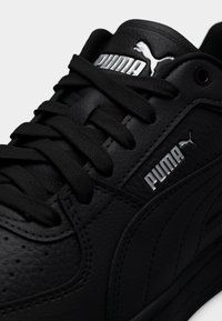 Μαύρα αθλητικά παπούτσια από υφή δέρματος με επίπεδα μαύρα κορδόνια. Διαθέτουν ασημένιο λογότυπο Puma και διακοσμητικά στοιχεία, με ένα ομαλό, στρογγυλεμένο σχέδιο.