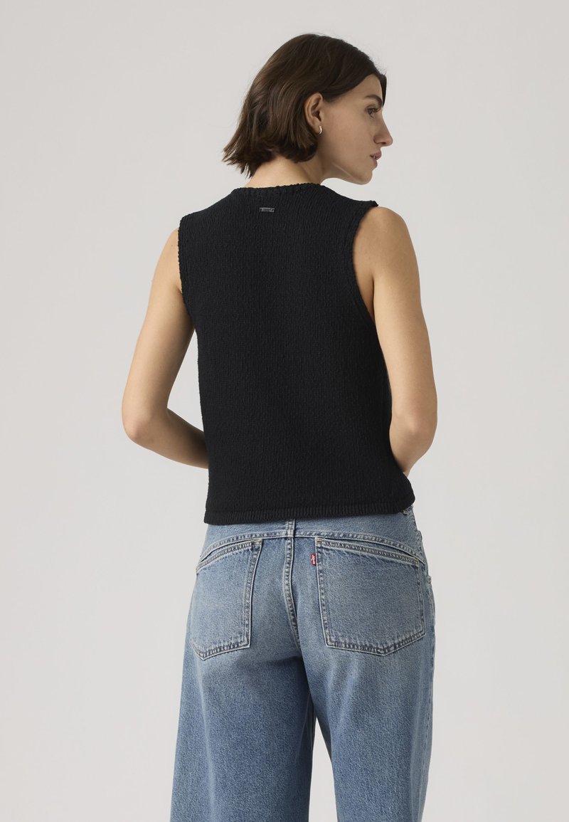 Woman with short brown hair wearing a sleeveless black top and light blue high-waisted jeans, facing away against a plain background.