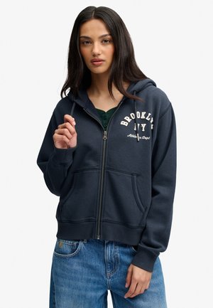 Femme aux longs cheveux foncés portant un sweat à capuche zippé bleu marine avec le texte "Brooklyn NY" et un jean bleu sur un fond uni.