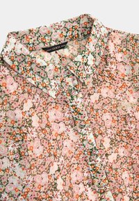 Bloemenblouse met een groen, roze en oranje patroon op een lichte, semi-transparante stof. Kraag en knoopdetails zichtbaar.