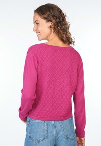 Fuchsia sweater with a textured knit pattern on the back, long sleeves, and a rounded neckline, paired with denim bottoms.