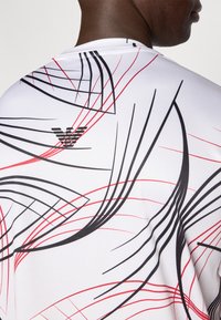 Λευκό αθλητικό μπλουζάκι με δυναμικό σχέδιο από κόκκινες και μαύρες γραμμές. Μαύρο λογότυπο  στο πάνω μέρος της πλάτης. Ομαλή υφή.