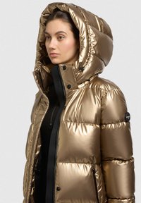 Auksinė pūkinė striukė su blizgiu paviršiumi, dideliu gobtuvu, užtrauktuku priekyje ir juodais akcentais, su logotipo detale ant rankovės.