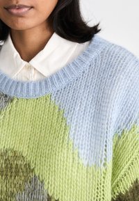 Pull en tricot avec un motif dans des teintes de bleu, vert et beige. Il a un col large et des poignets côtelés. Le fil texturé ajoute de la profondeur.