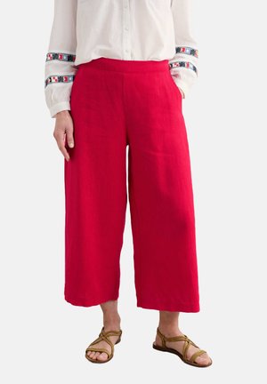 Pantaloni cropped rossi in lino con design a gamba ampia, texture liscia e tasche laterali, abbinati a una camicia bianca con bottoni e dettagli decorativi.