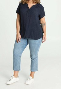 Femme portant un chemisier bleu marine, un jean court bleu clair, des baskets blanches, avec un tatouage floral sur son avant-bras gauche.