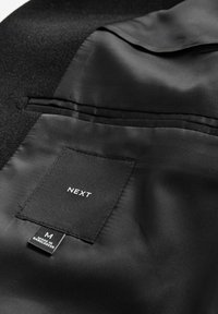 Interiorul unui blazer negru, cu o căptușeală din satin neted și un buzunar, etichetat "NEXT". Eticheta de îngrijire indică mărimea "M" și originea "FABRICAT ÎN BANGLADEȘ".