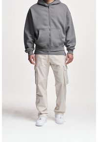 Šedá mikina na zip s předními kapsami, ve spojení s béžovými cargo kalhotami s bočními kapsami a bílými teniskami. Jednoduchý, ležérní outfit.