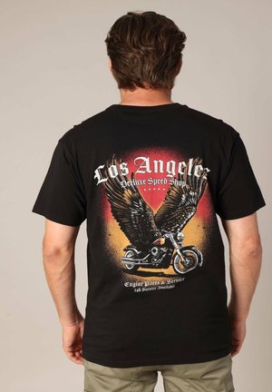 Zwarte katoenen t-shirt met een afbeelding van een motorfiets en grote vleugels, met een achtergrond in rood en oranje, en tekst die luidt: "Los Angeles Deluxe Speed Shop."