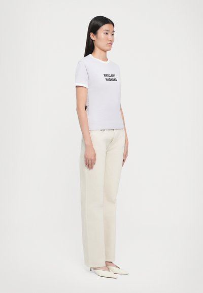 Tricou alb din bumbac cu text negru "BRILLIANT MADNESS", asociat cu pantaloni larga de culoare crem și pantofi cu vârf ascuțit.