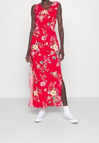 Punainen kukkakuviollinen maxi-mekko, jossa on hihattomuus, sivuhaalari ja naru vyötäröllä, yhdistetty valkoisiin tennareihin. Mekossa on keltaisia ja vihreitä yksityiskohtia.