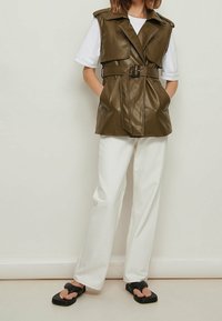 Olivenfarbener Kunstlederwesten mit einem Gürtel, Reverskragen und frontalen Taschen, kombiniert mit weißen, weit geschnittenen Hosen und schwarzen flachen Sandalen.