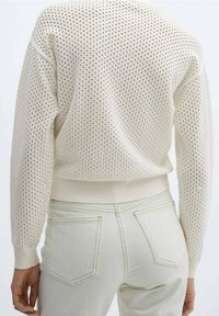 Light beige crochet sweater with a textured, perforated design and ribbed hem, paired with pale jeans featuring back pockets.