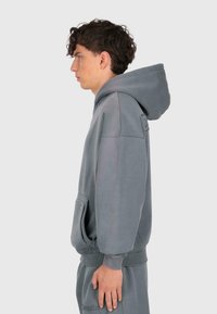 Junger Mann mit lockigem Haar, der einen übergroßen grauen Kapuzenpullover und passende Hose trägt, seitlich vor einem schlichten Hintergrund stehend.
