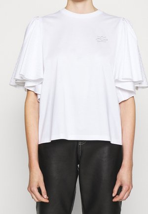 Vrouw die een wit topje draagt met korte mouwen met gelaagde ruches en een zwarte broek met zichtbare witte stiksels.