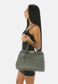 Bolso de cuero gris con un diseño estructurado, asa superior, herrajes dorados y una etiqueta desmontable. Superficie texturizada y acentos metálicos.